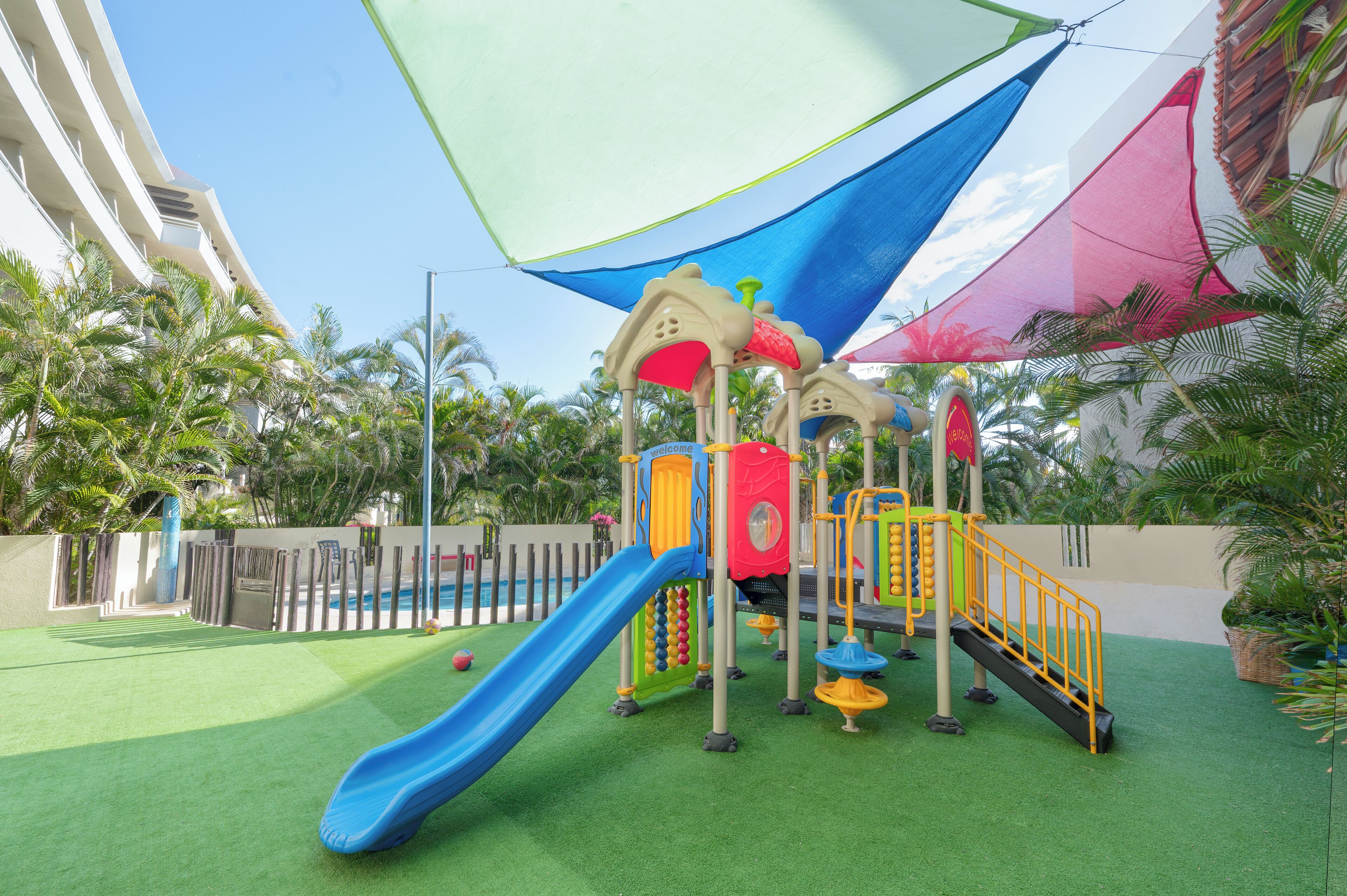 Área de recreação infantil com escorrega e estruturas de escalada, cercada por palmeiras e uma piscina.