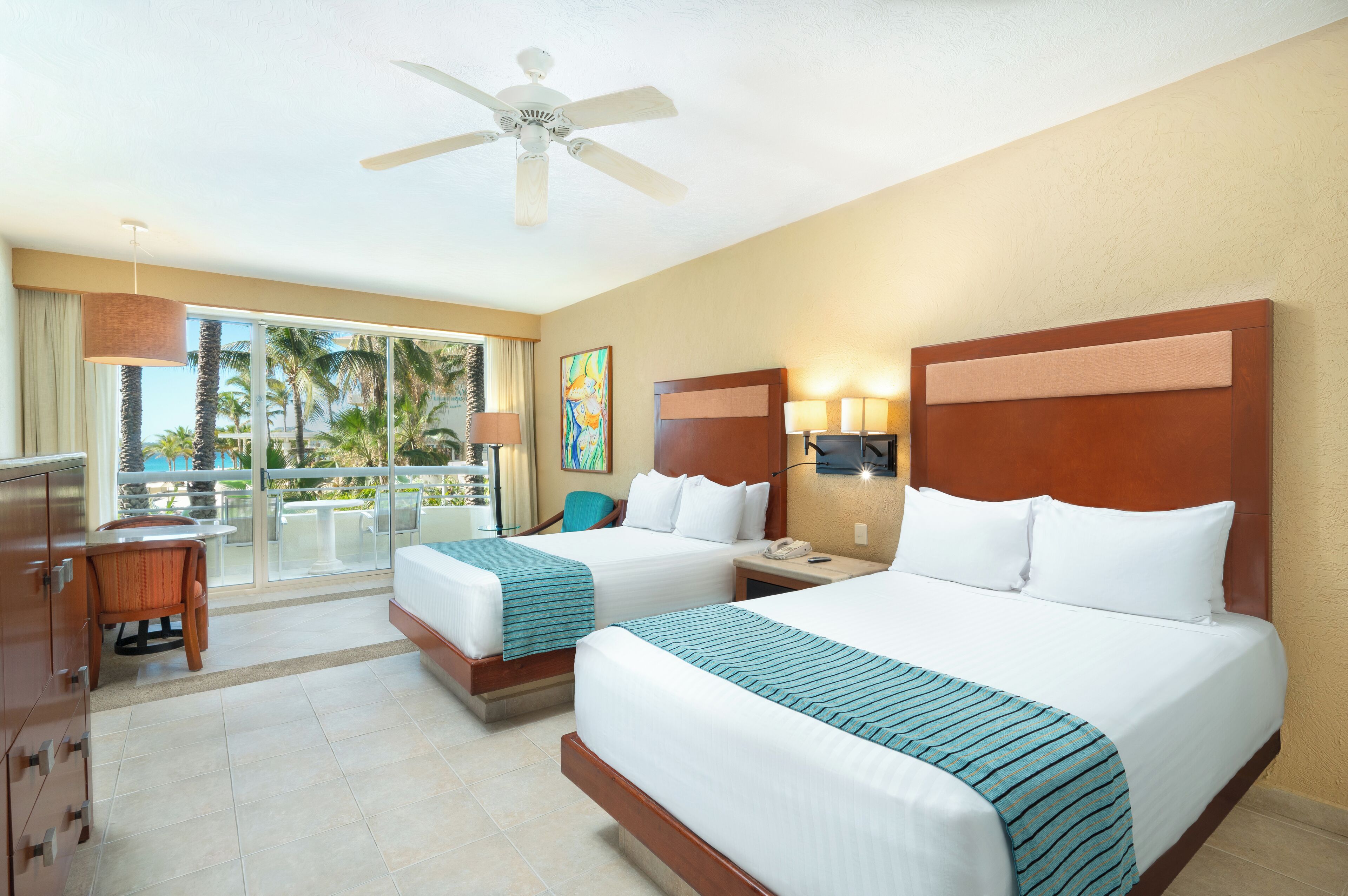 A hotel room with two beds, a desk, a chair, a balcony with a view of palm trees, and a ceiling fan.