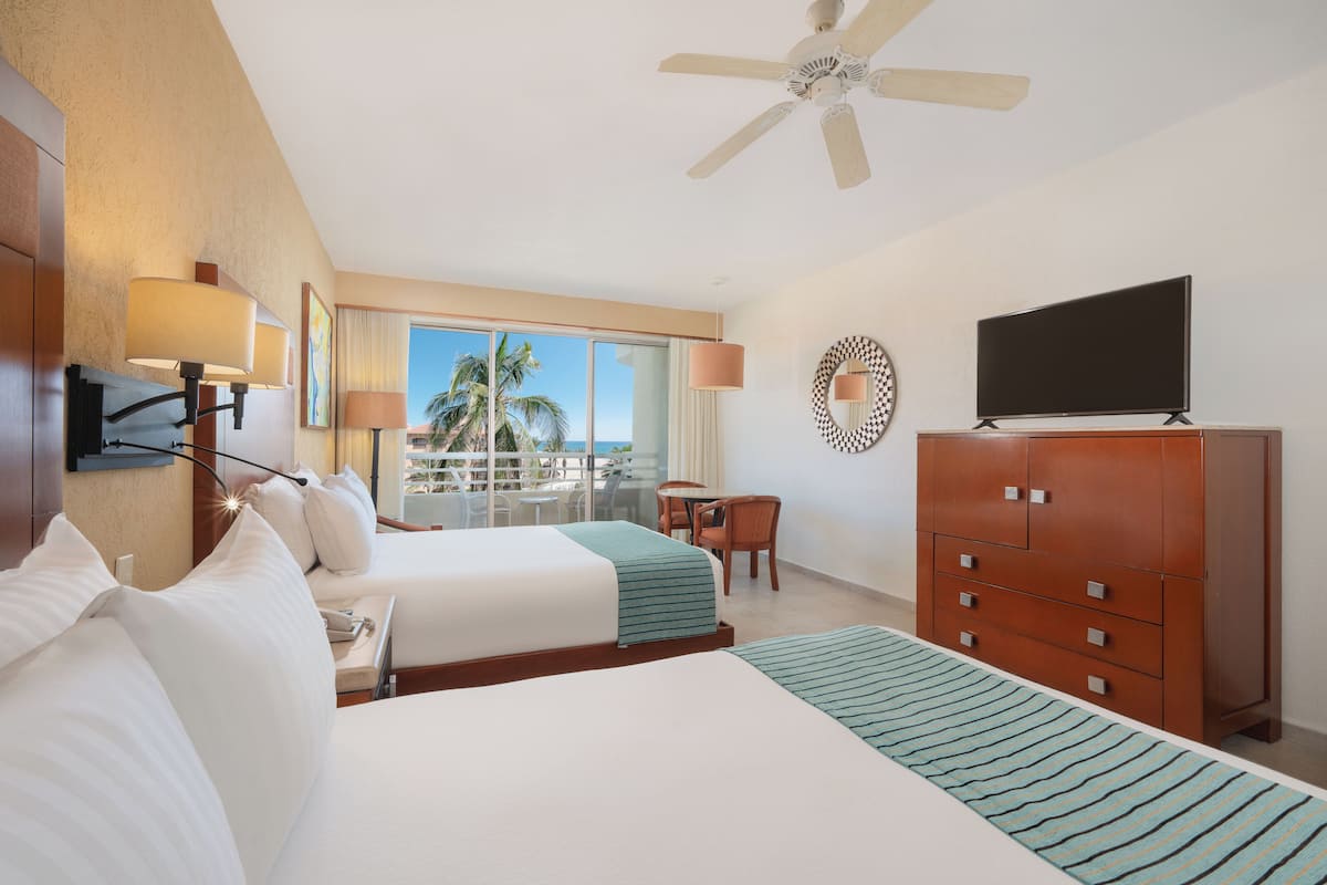 A hotel room with two beds, a TV, a ceiling fan, a desk, and a view of palm trees.