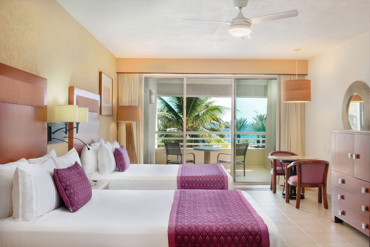 A hotel room with two beds, a dining area, and a balcony with a view of palm trees.