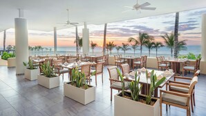 A spacious outdoor dining area with multiple tables and chairs, large windows offering a view of the sunset over the ocean, and ceiling fans. - GR Solaris Lighthouse Los Cabos (San José del Cabo)