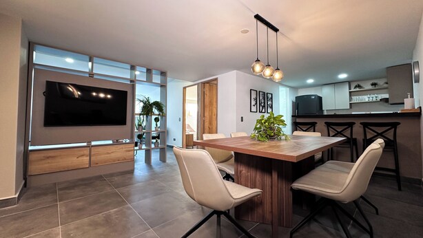 A modern living room with a large flat-screen TV, a wooden dining table with four chairs, and a kitchen area with a TV and counter space. - Amplio apt en poblado by Hoost APT 701 (Medellín)