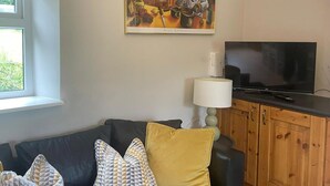 A living room with a dark-colored sofa, patterned cushions, a painting above the sofa, a television on a wooden cabinet, and a window with a view of greenery. - Cottage in the Dublin Mountain (Cunard)