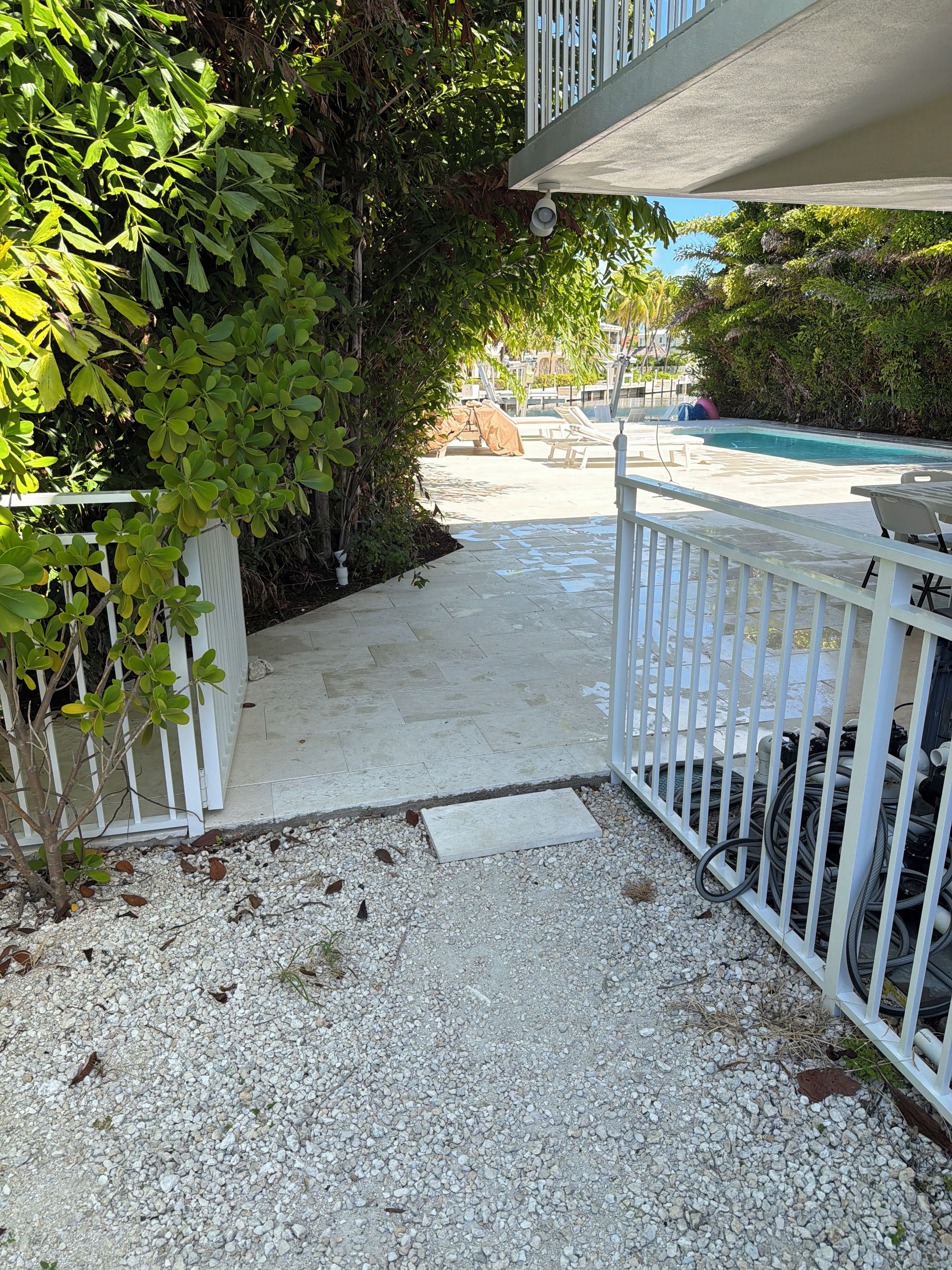 A pathway leading to a pool area with a white fence, gravel ground, and greenery.