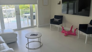 Ein modernes Wohnzimmer mit einem weißen Sofa, einem runden Couchtisch und einem an der Wand montierten Fernseher. Durch die Tür ist ein Balkon mit Geländer zu sehen.