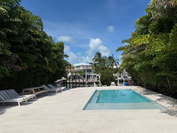 A swimming pool with lounge chairs, surrounded by dense foliage and a building in the background.