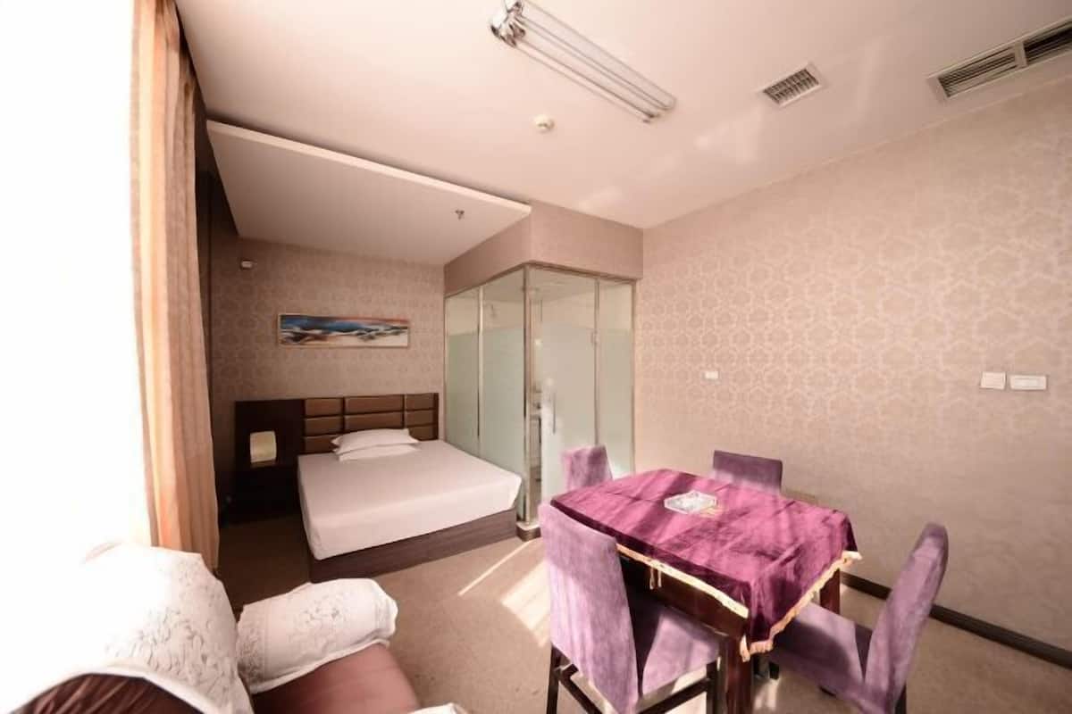 A hotel room with a bed, a glass shower enclosure, a dining table with purple chairs, and a painting on the wall.