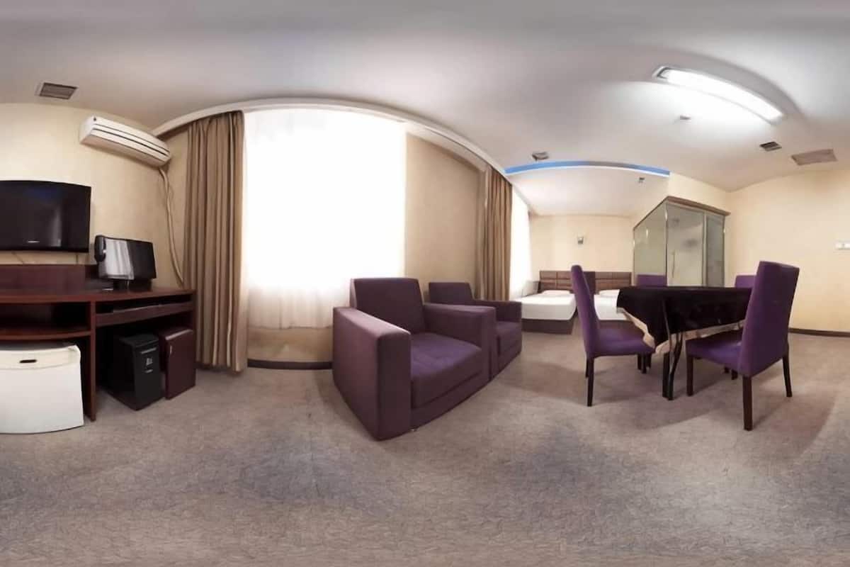 A hotel room with a flat-screen TV, a desk with a computer, a sofa, a dining table with chairs, and a large window with curtains.