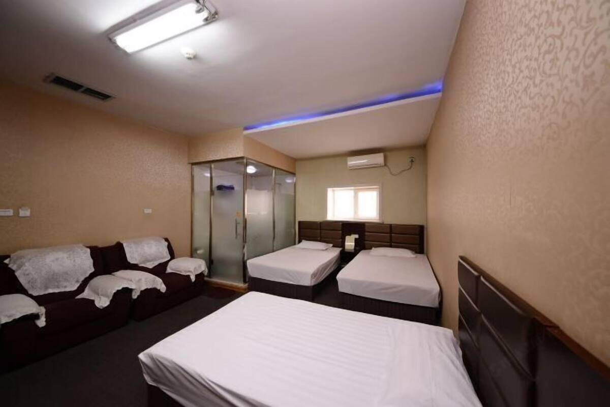A hotel room with two single beds, a sofa, a glass-enclosed shower, and a window.
