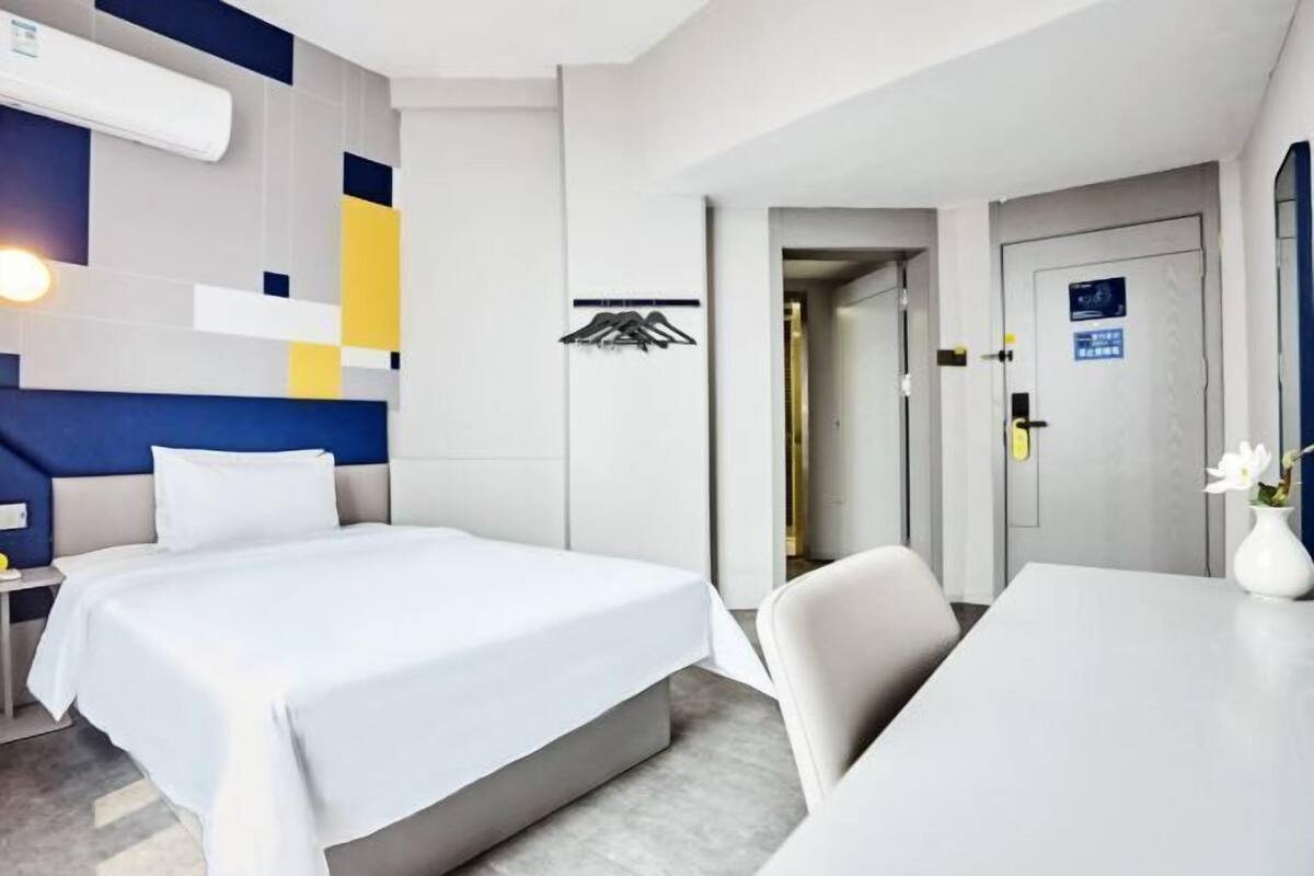 A hotel room with a bed, a desk, and a chair. There is a door with a sign on it.