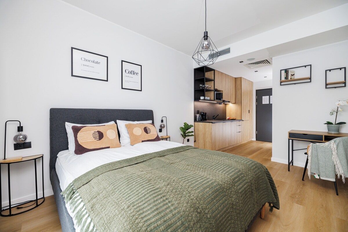 Een moderne slaapkamer met een groot bed, een nachtkastje, een bureau en een keuken in de achtergrond.