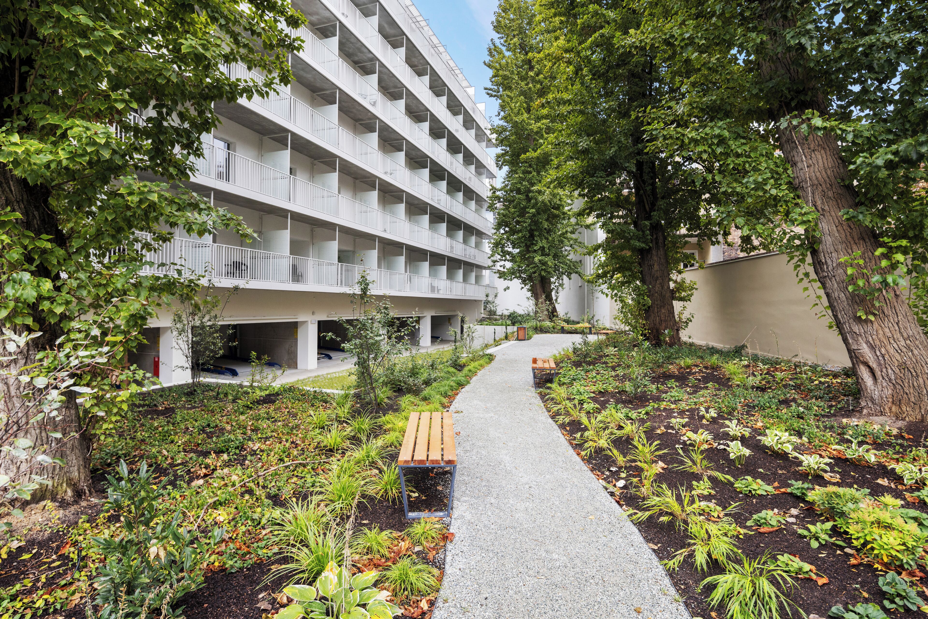 Un immeuble à étages, doté de balcons, entouré d’un jardin aménagé avec un chemin de gravier et des bancs.