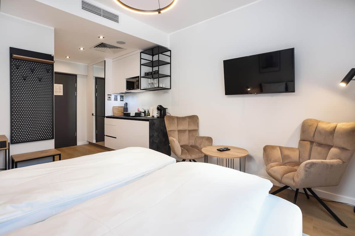 Una habitación de hotel moderna con una cama grande, dos sillones, una pequeña mesa redonda, un televisor montado en la pared, una zona de cocina y un estante negro original.