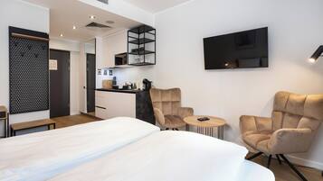 A modern hotel room with a large bed, two armchairs, a small round table, a TV mounted on the wall, a kitchen area, and a unique black shelving unit.