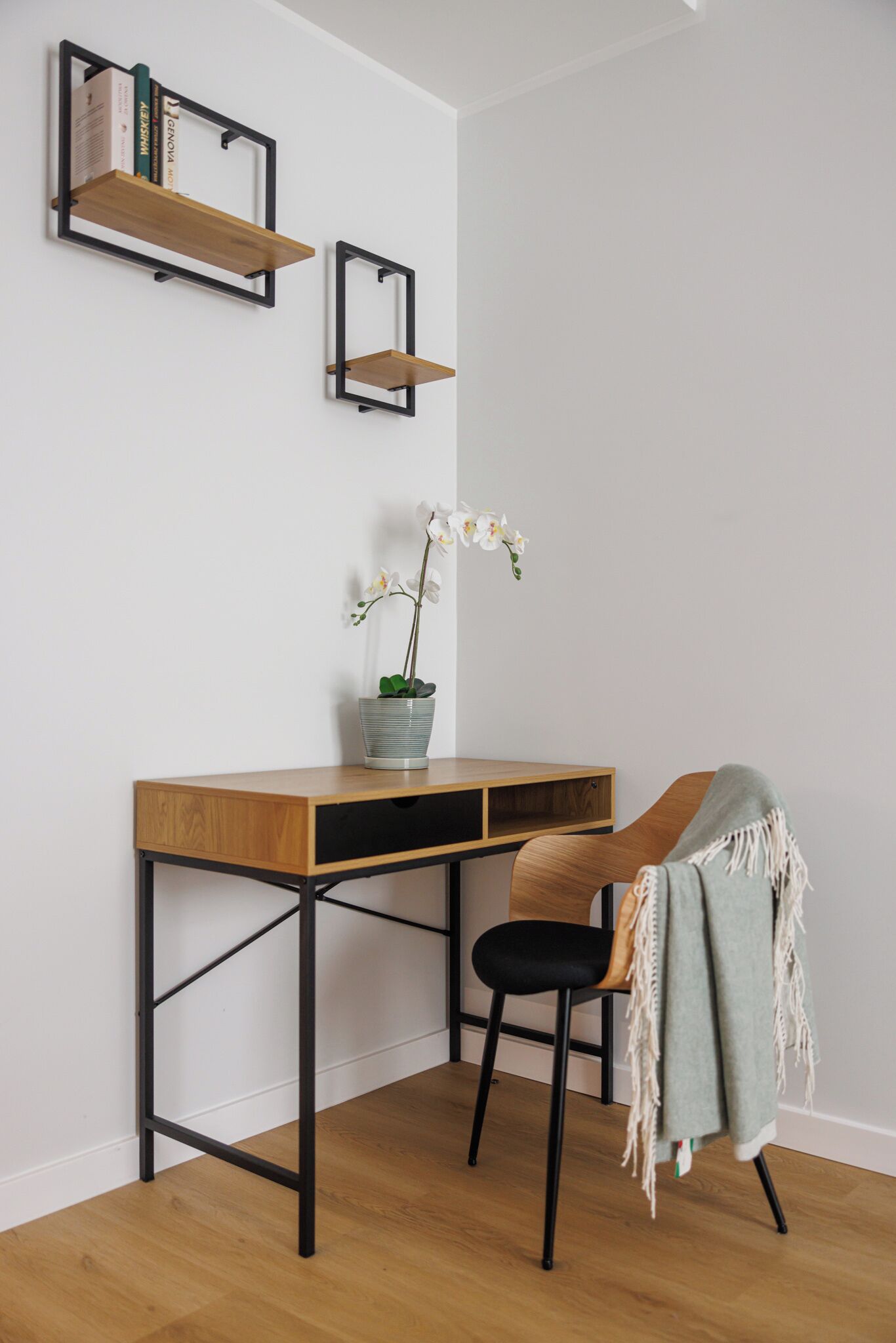 A corner desk with a chair, a potted orchid, and two wall-mounted shelves with books.