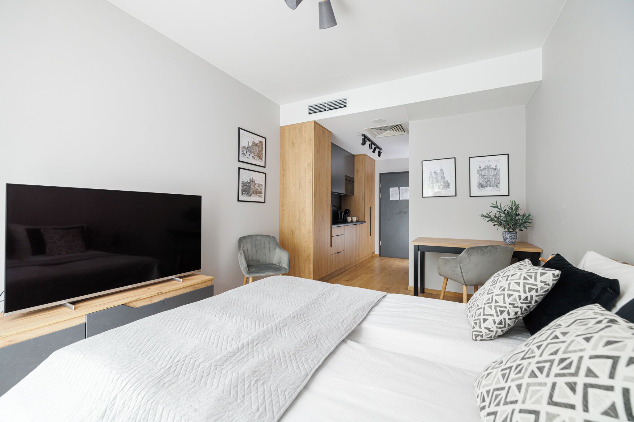 A modern bedroom with a large bed, a television, a desk, and a kitchenette in the background.