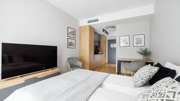 A modern bedroom with a large bed, a television, a desk, and a kitchenette in the background.