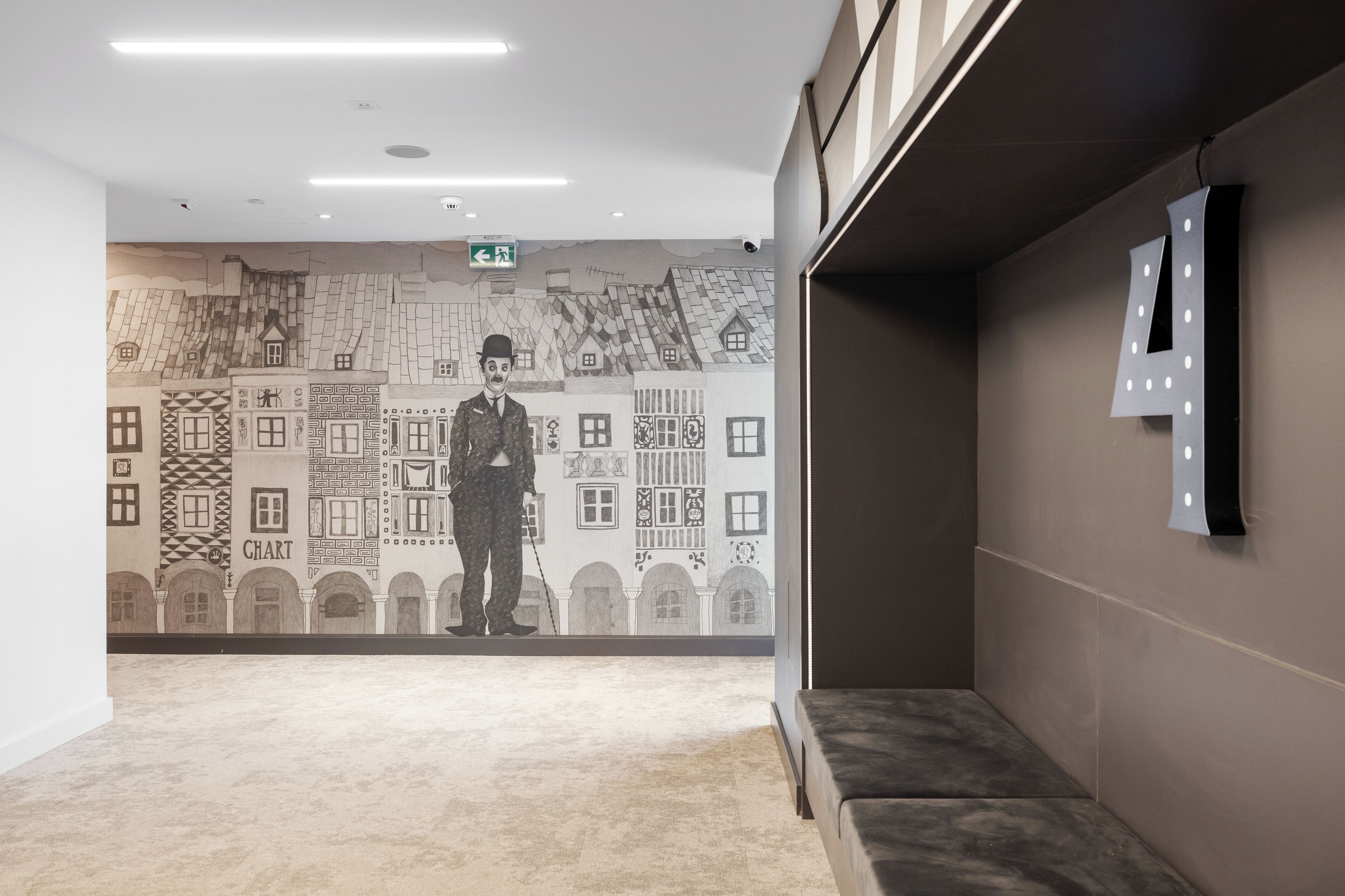 A hallway with a mural of buildings and a person standing in the center. There is a bench on the right and a wall-mounted light fixture.