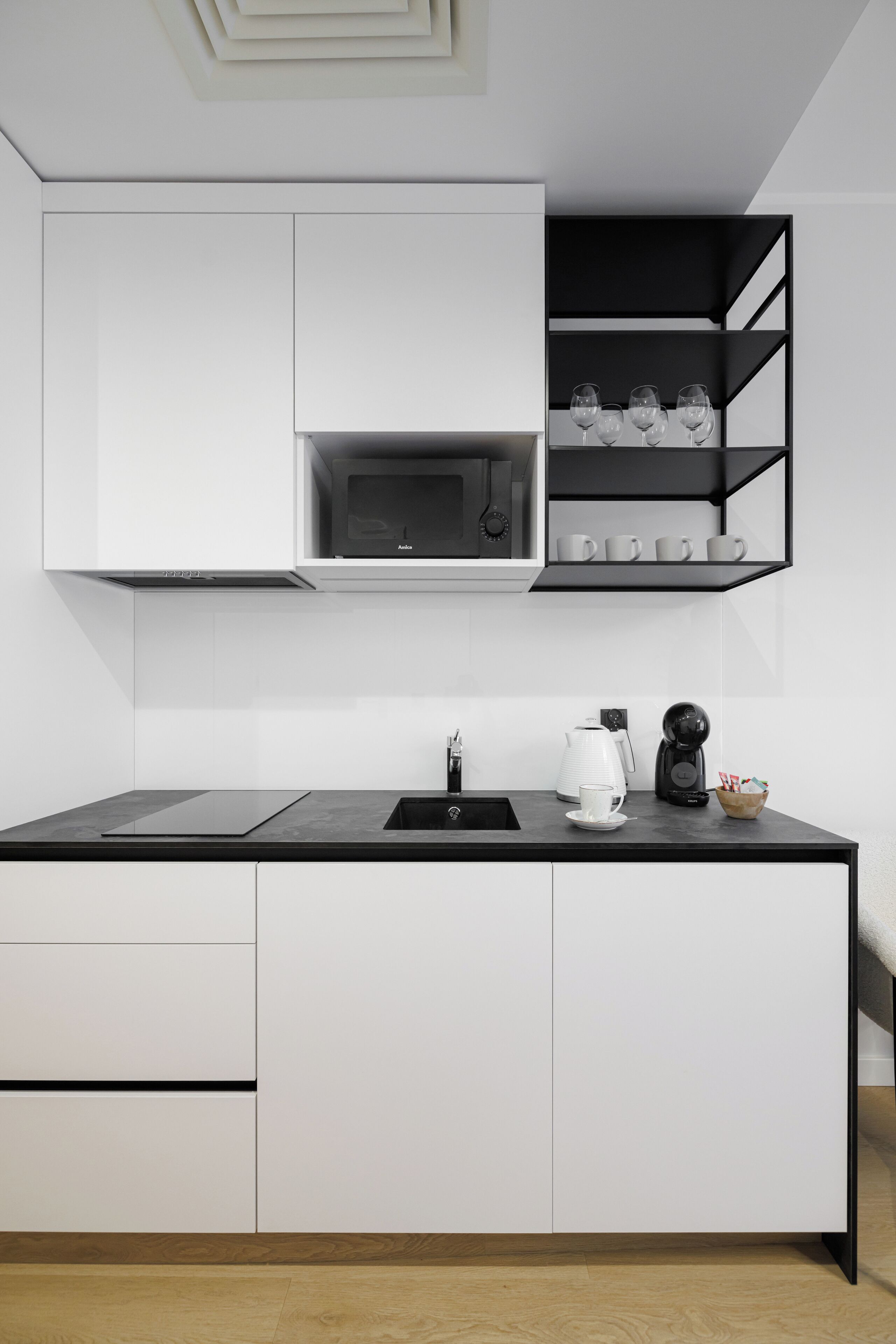 A modern kitchen with white cabinets, a black countertop, a microwave, a coffee maker, and a shelf with glasses.