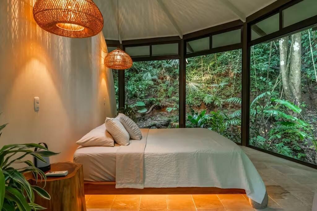 Une chambre à coucher avec un lit, un luminaire suspendu et une vue sur une forêt, visible à travers de grandes fenêtres.