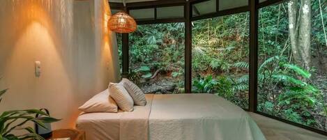Une chambre à coucher avec un lit, un luminaire suspendu et une vue sur une forêt, visible à travers de grandes fenêtres.