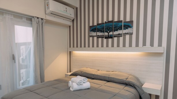 A neatly made bed with a grey duvet and pillows, a set of folded towels, a wall-mounted air conditioner, and a striped wall with a framed picture above the bed. - Maison Work & Travel 2 - Palermo  (Buenos Aires)