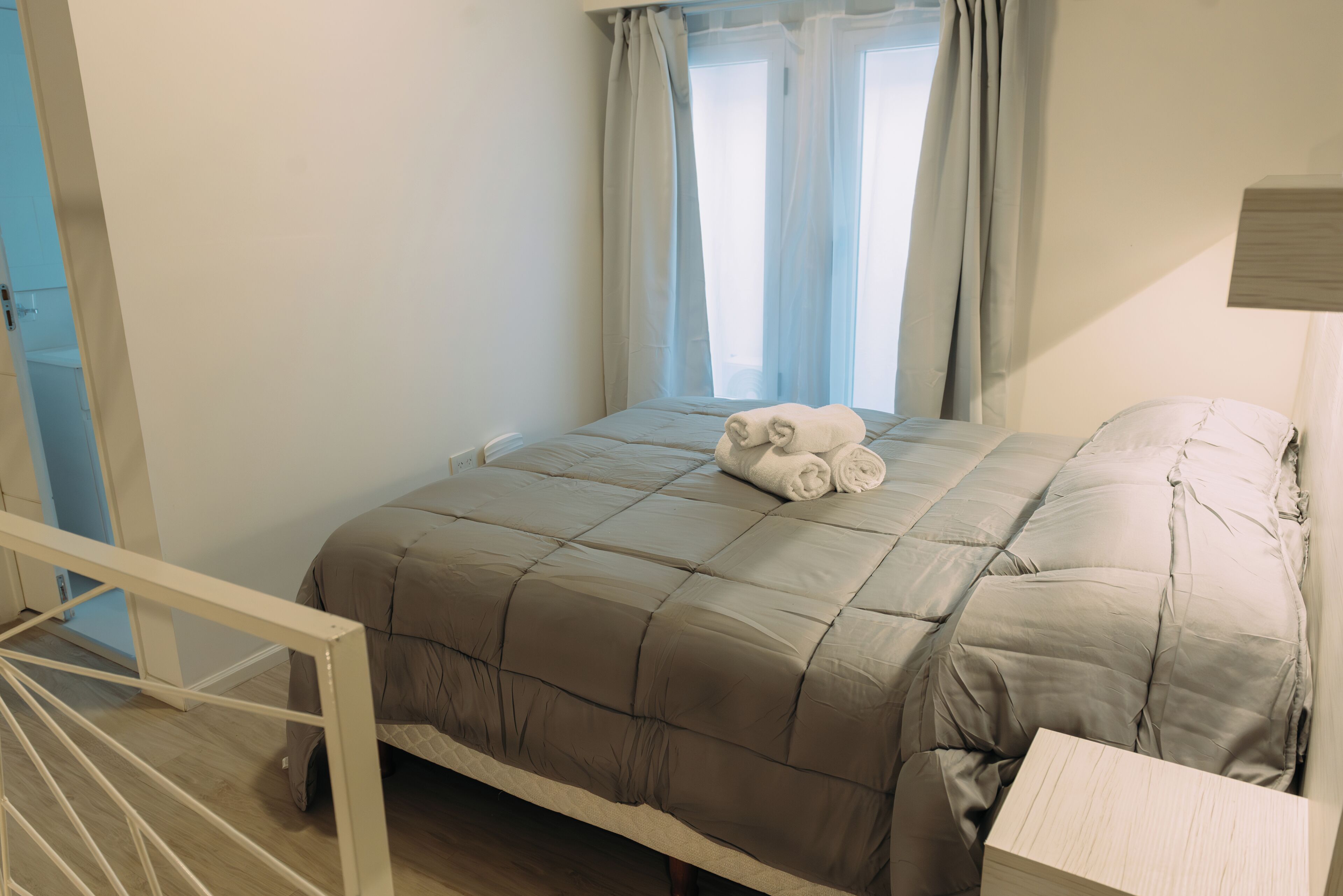 Una cama bien hecha con un edredón color beige, una mesita de noche de madera y una ventana con cortinas azul claro.