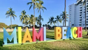 Un letrero con las palabras 'MIAMI BEACH' en letras coloridas sobre un área verde con palmeras y un edificio al fondo.