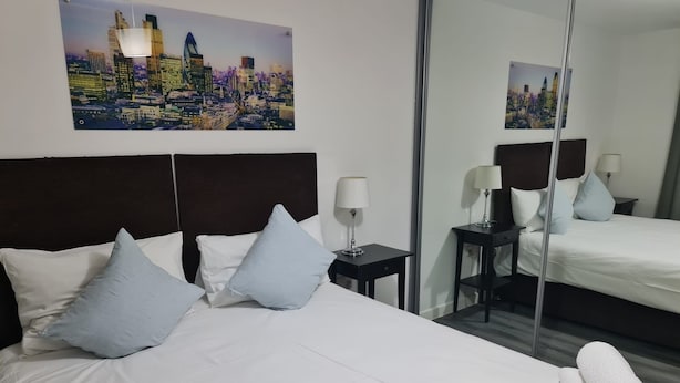 A hotel room with a large bed, two bedside tables with lamps, a mirror, and a cityscape painting on the wall. - Stratford Luxury Apartments (London)