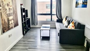 A modern living room with a grey sofa, a coffee table, and a bookshelf. A large window with grey curtains allows natural light to enter. - Stratford Luxury Apartments (London)