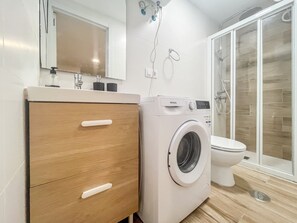 Ein modernes Badezimmer mit integrierter Waschmaschine, einem Holzkabinet mit Schubladen, einer Toilette und einer Duschkabine aus Glas.