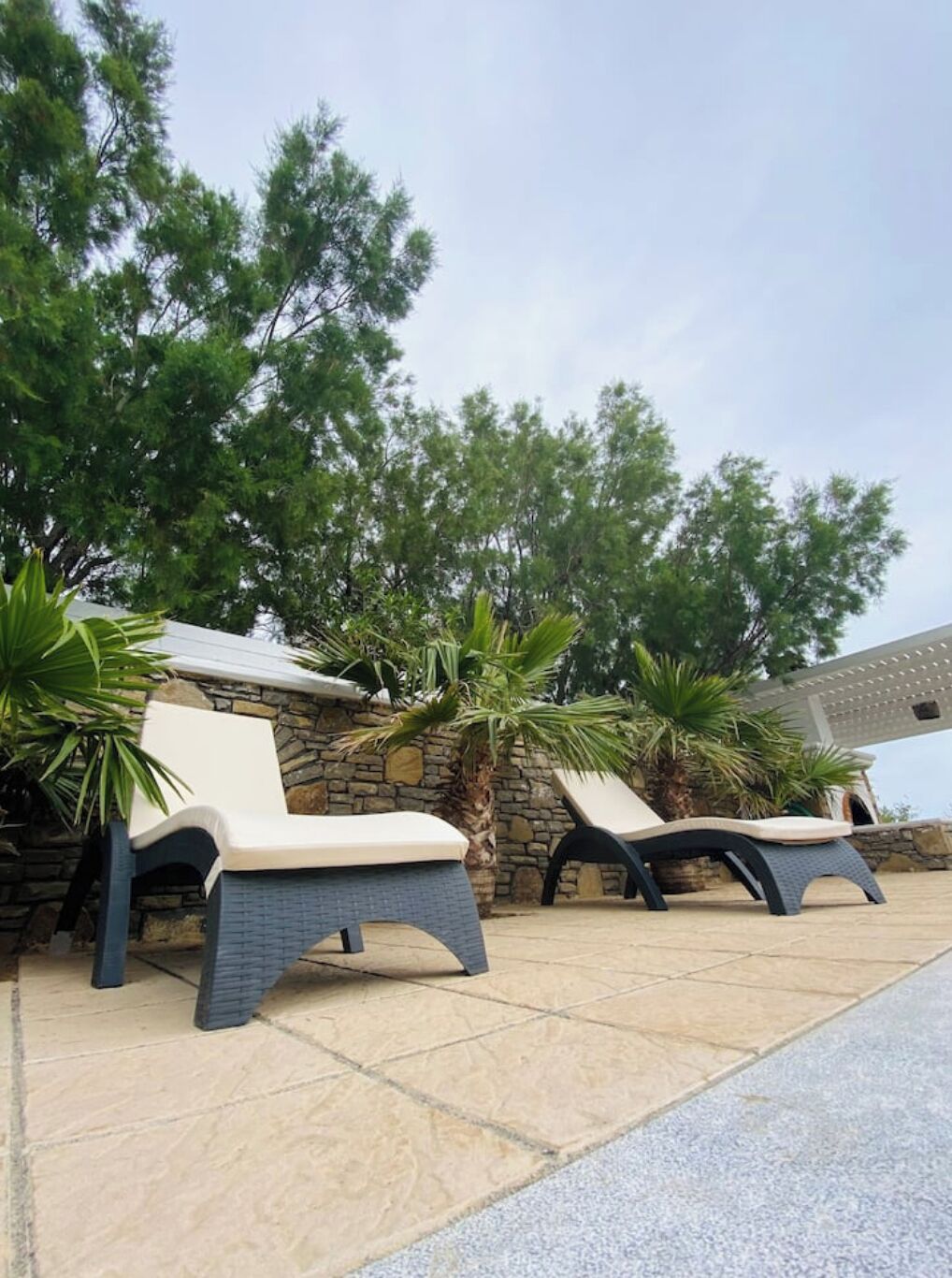 Two lounge chairs with cushions, a stone wall, and a pergola with a roof.