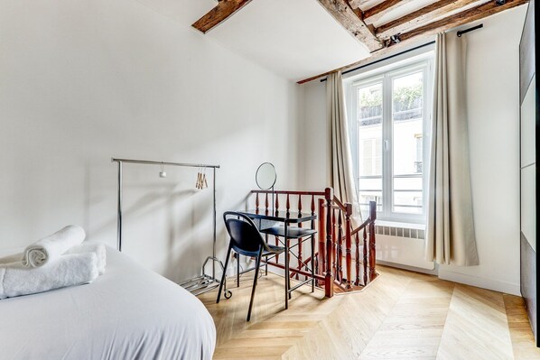 A bedroom with a bed, a desk with a chair, a window with curtains, and a wooden ceiling.