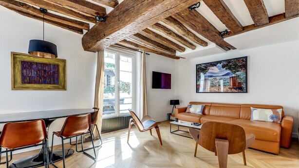 A modern living room with exposed wooden beams, a dining area with a table and chairs, and a large window providing natural light.