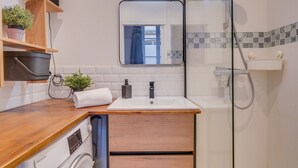 Een compacte badkamer met een houten aanrecht, een wasmachine, een spiegel en een douchehoek.