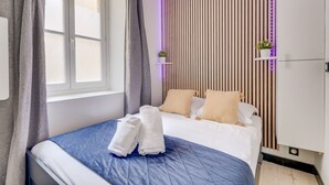 Een net opgemaakt bed met een blauwe quilt en witte handdoeken, een houten lambrisering met een kleine plant, en een witte inbouwkast.