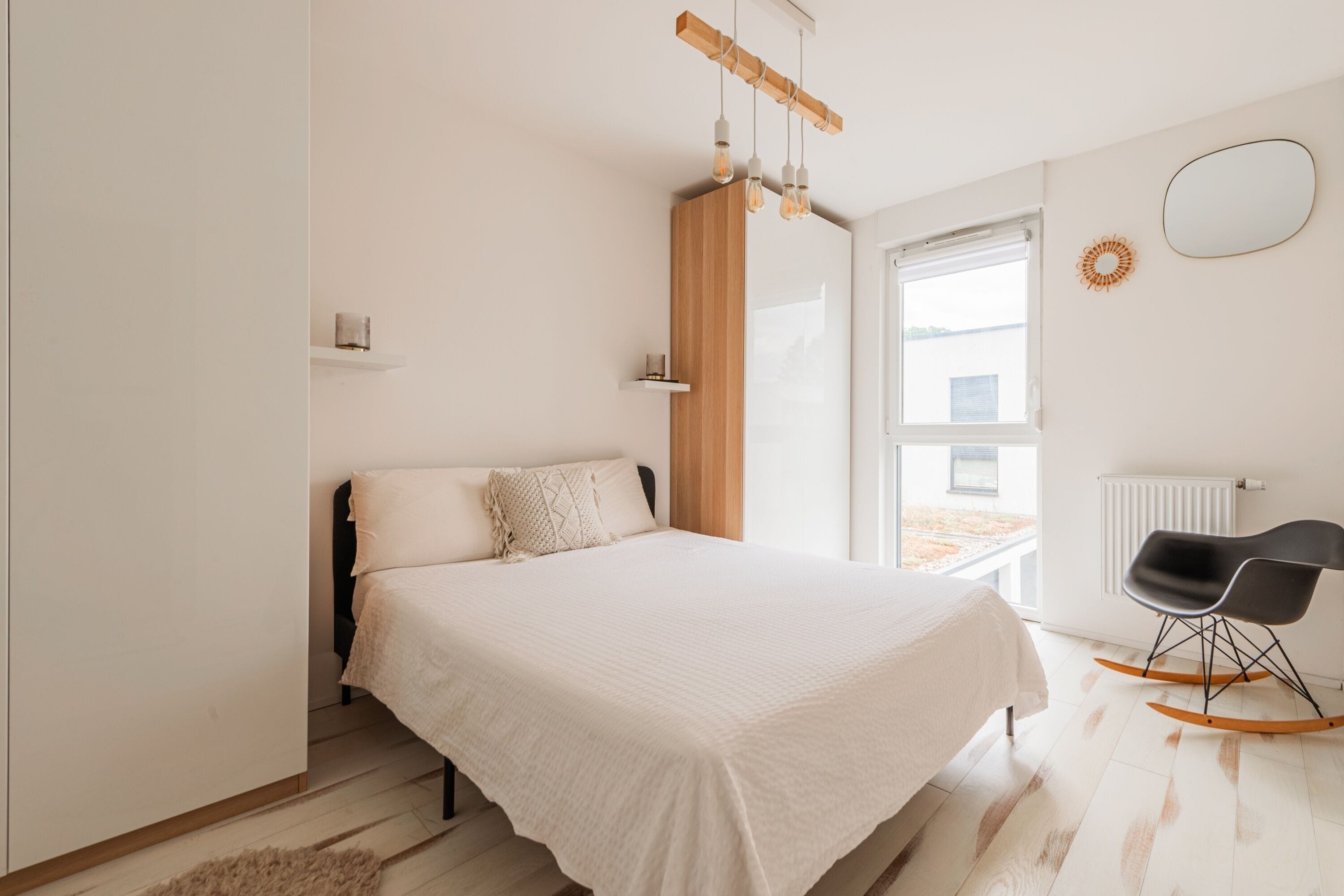 A bedroom with a bed, a wooden headboard, a window, a radiator, and a black rocking chair.