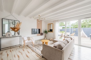 Ein modernes Wohnzimmer mit Sofa, Couchtisch und Fernseher. Ein großes Fenster bietet Blick ins Freie.