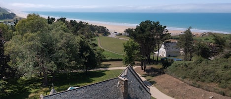 A coastal view with a beach, a pathway leading to a building with a stone roof, and a clear sky.