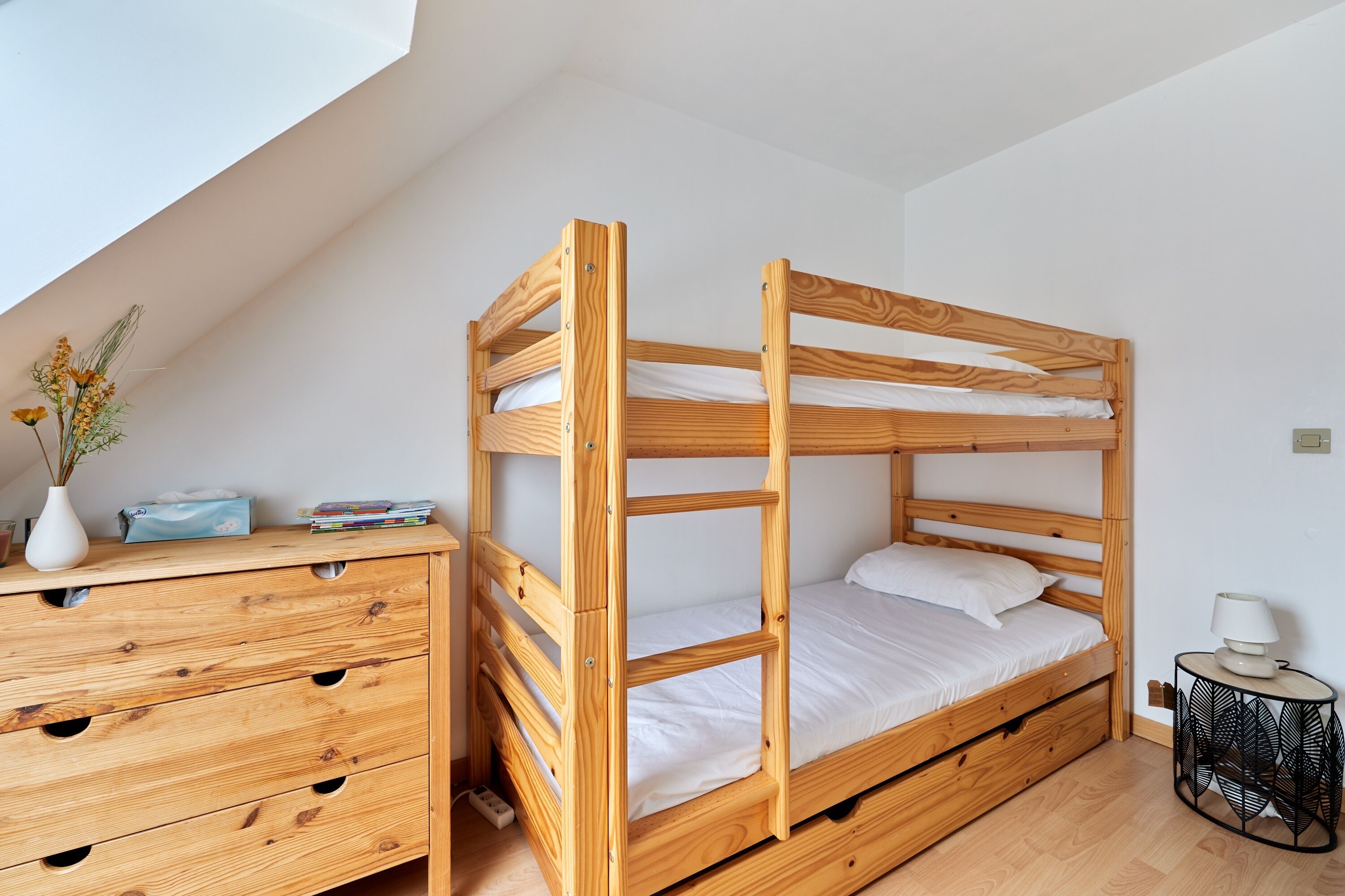 Ein Etagenbett mit Holtzargen und weißer Bettwäsche, eine Holzkommode mit mehreren Schubladen und ein kleiner runder Tisch mit einer Lampe.
