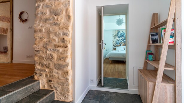 Une pièce avec un mur en pierre, un escalier en bois, un lit avec une tête de lit bleue et une étagère en bois où sont posés des livres et une plante.