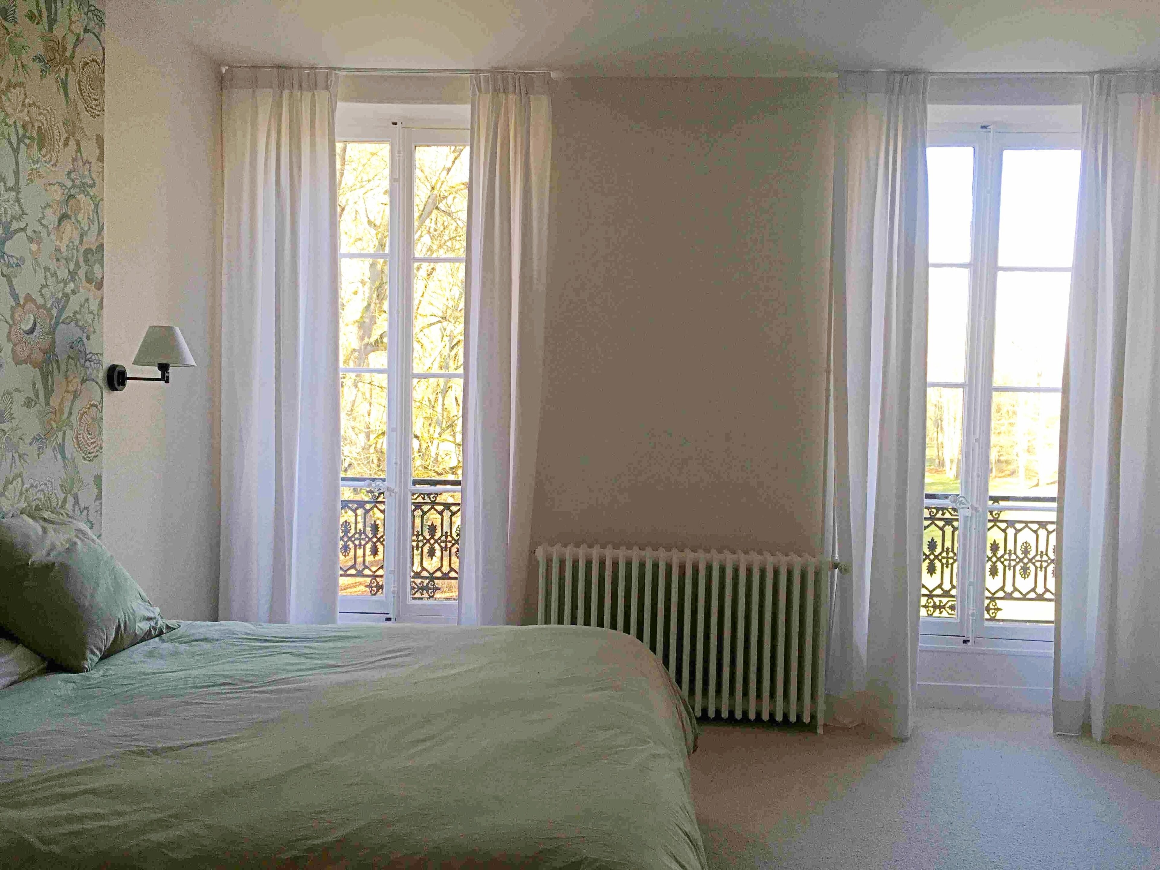 Een slaapkamer met een bed, twee ramen met gordijnen, een radiator en een wandlamp.