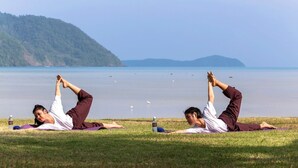 Twee personen doen yoga op grasmatten aan een meer, met bergen op de achtergrond.