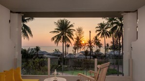 A balcony with a yellow sofa and a wooden rocking chair, overlooking a beach with palm trees and a sunset.
