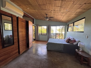 Quarto com cama, ventoinha no teto, janela e porta que dá para uma varanda.