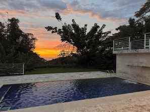 Piscina com vista para o pôr do sol, cercada por árvores e uma estrutura de concreto.
