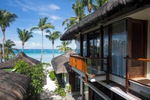 Un hotel frente a la playa con balcón con vista al océano, palmeras y construcciones de techo de paja.