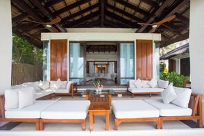 A covered outdoor seating area with white sofas and wooden tables, a central coffee table, and a view of the interior through glass doors. - Harlan Beach Villa Boracay (Boracay Island)
