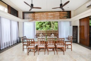Un comedor con una mesa de vidrio rodeada de sillas, ventiladores de techo y vistas a zonas verdes exteriores.
