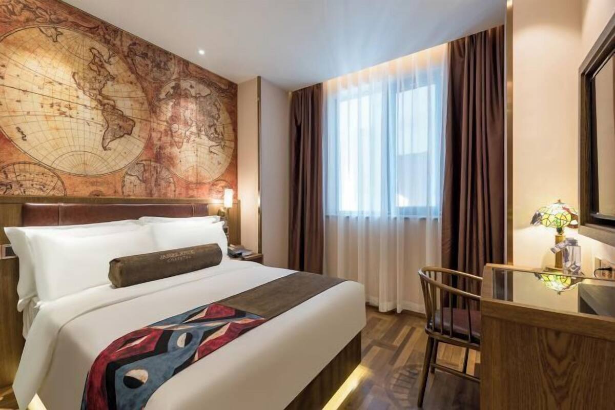 Una habitación de hotel con un gran mapa mundial en la pared, una cama con una colcha estampada, un escritorio con una lámpara y vistas al exterior a través de una ventana con cortinas.